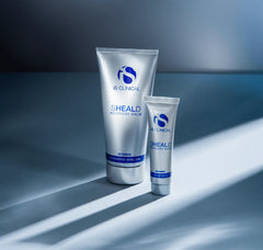 Two tubes of IS Clinical SHEALD Recovery Balm on a gray surface with a gradient background.